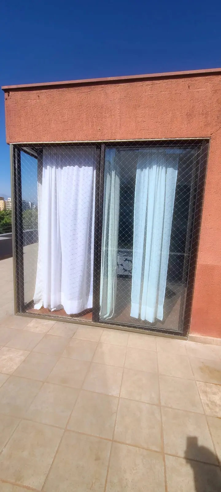 Ventanal corredero de departamento cerrado con malla de seguridad, visto desde una terraza con piso de cerámica y muro terracota